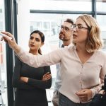 Women’s Experiences in the Workplace: Progress Without Real Change Woman, group and collaboration with notes on board with review, listening and planning with brainstorming. Coach, training and team by glass wall, questions and discussion with strategy in office.